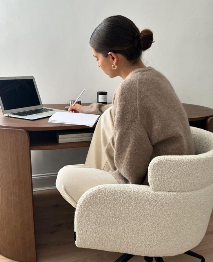 Creative at a curved wood desk in a bouclé chair, writing in a notebook beside an open laptop — bright, neutral studio.