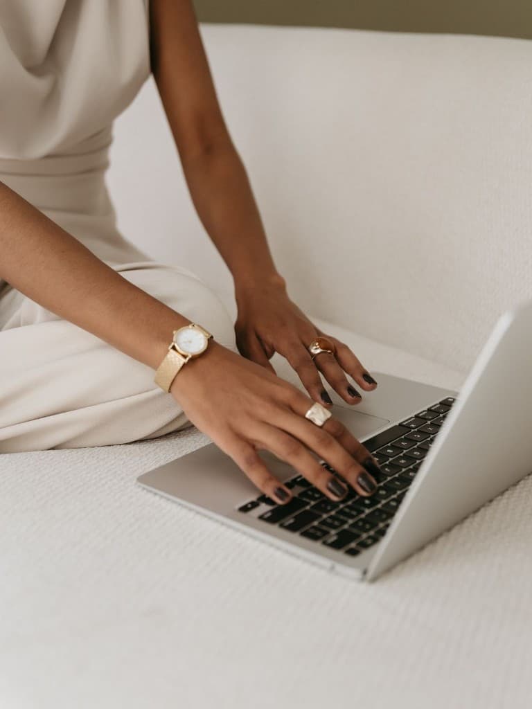 Close-up of hands typing on a silver laptop, cream loungewear, gold watch and rings, soft bright background — calm professional mood.